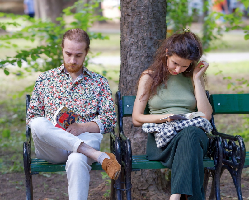 15 - Two individuals reading books while sitting on a bench in a city park